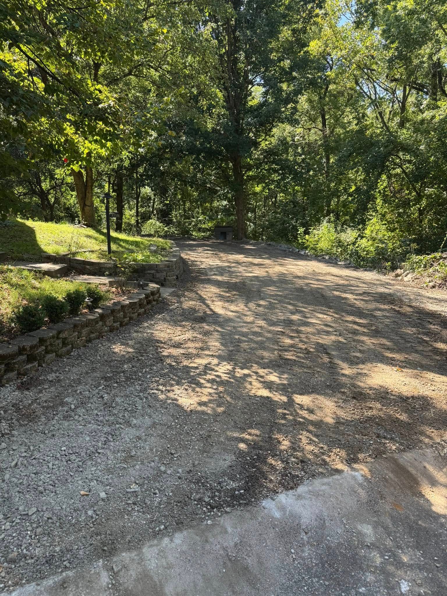 Gravel road leads into a wooded area, sunlight filtering through trees.