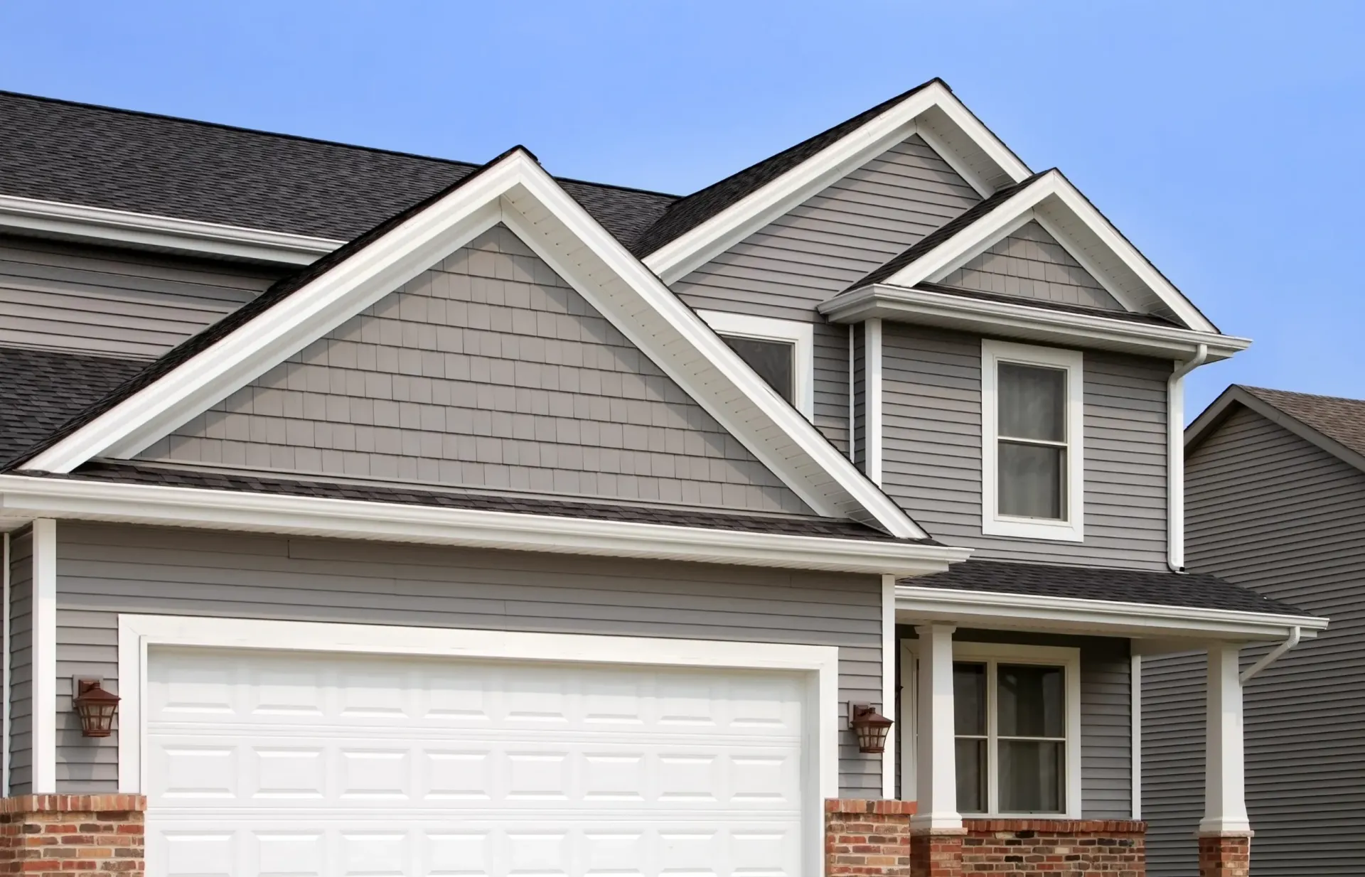 A close-up view of the roof, gray siding, and white garage door of a modern suburban house against a blue sky.
