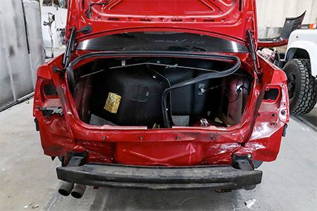 Red car trunk open, damaged rear. Shows trunk interior and exposed components in repair shop.