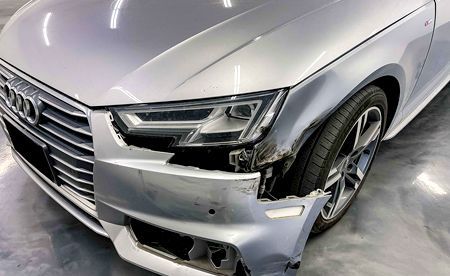 Damaged silver Audi car front, bumper detached, headlight intact, showing a side view.