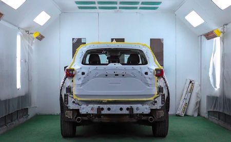 Rear view of a partially assembled white car inside a paint booth with lights and masking tape.