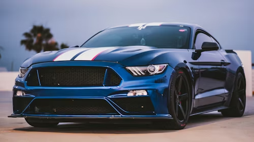 Blue sports coupe parked outdoors at dusk, front view with black accents and blurred palm trees in the background