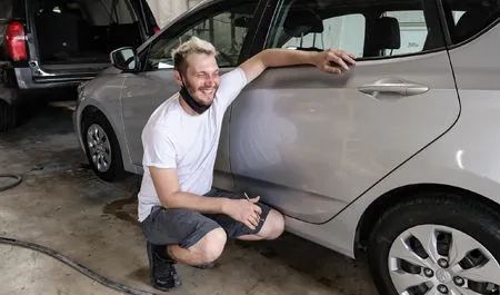 Man kneeling beside a silver car in a garage, smiling and pointing at the side panel