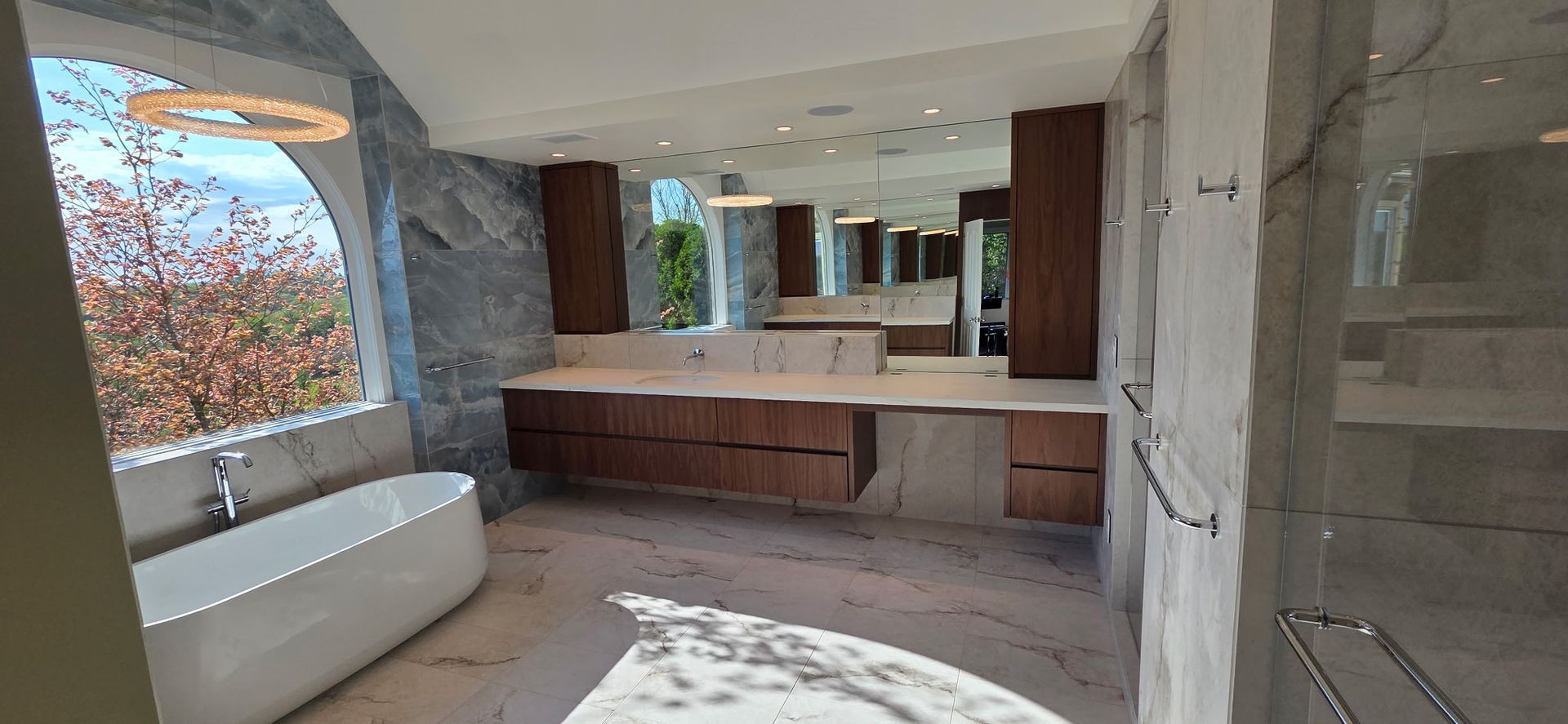 Modern bathroom with double vanity, large mirror, marble walls, and a white bathtub by the window