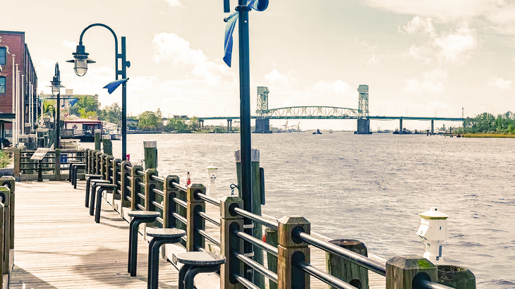 Waterfront walkway with benches, lampposts, and a bridge in the distance.