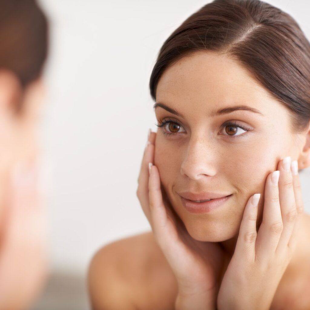 Woman looks at her reflection in a mirror, smiling. She touches her face with both hands, light-skinned.