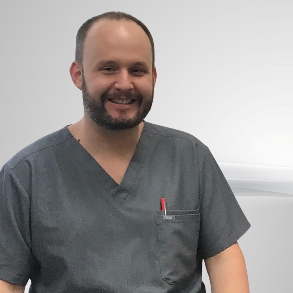 Man with a beard wearing gray scrubs, smiling, and a pen in his pocket.
