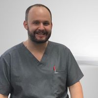 Man with a beard wearing gray scrubs, smiling, and a pen in his pocket.
