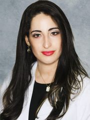 Woman with long dark hair, red lipstick, wearing a white lab coat.