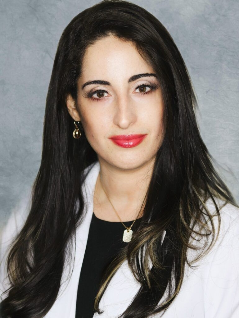 Woman with long dark hair, red lipstick, wearing a white lab coat.