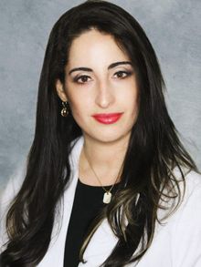 Woman with long dark hair, red lipstick, wearing a white lab coat.