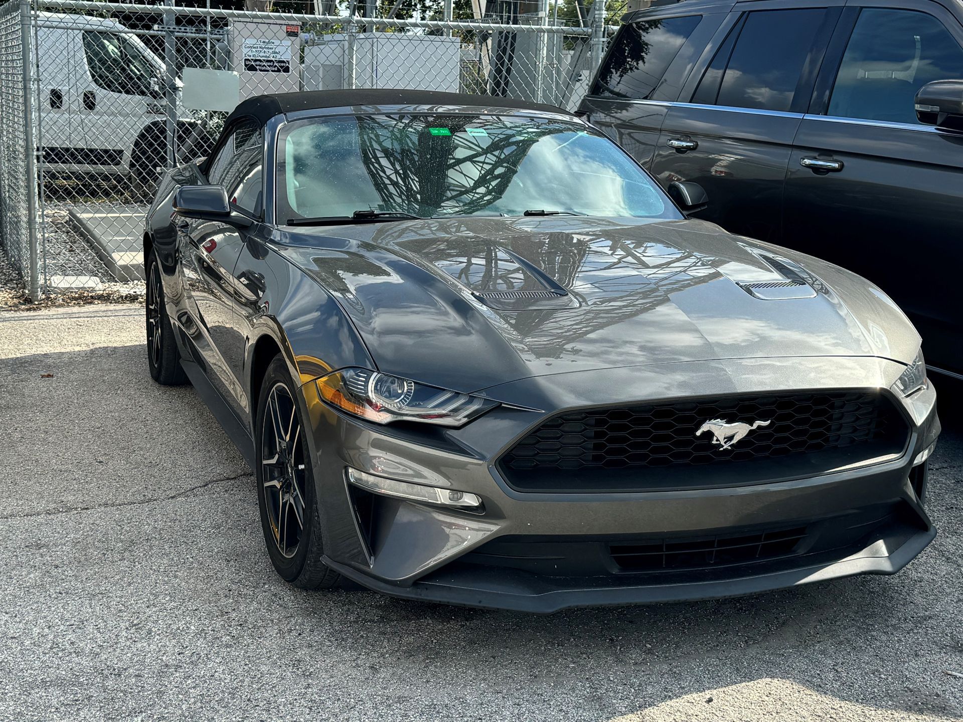 A gray mustang convertible is parked in a parking lot next to a fence.