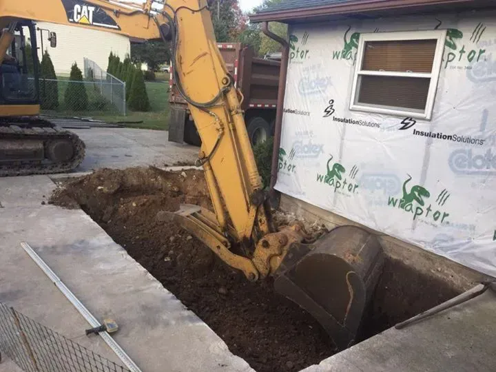 A cat excavator is digging a hole in the ground in front of a house