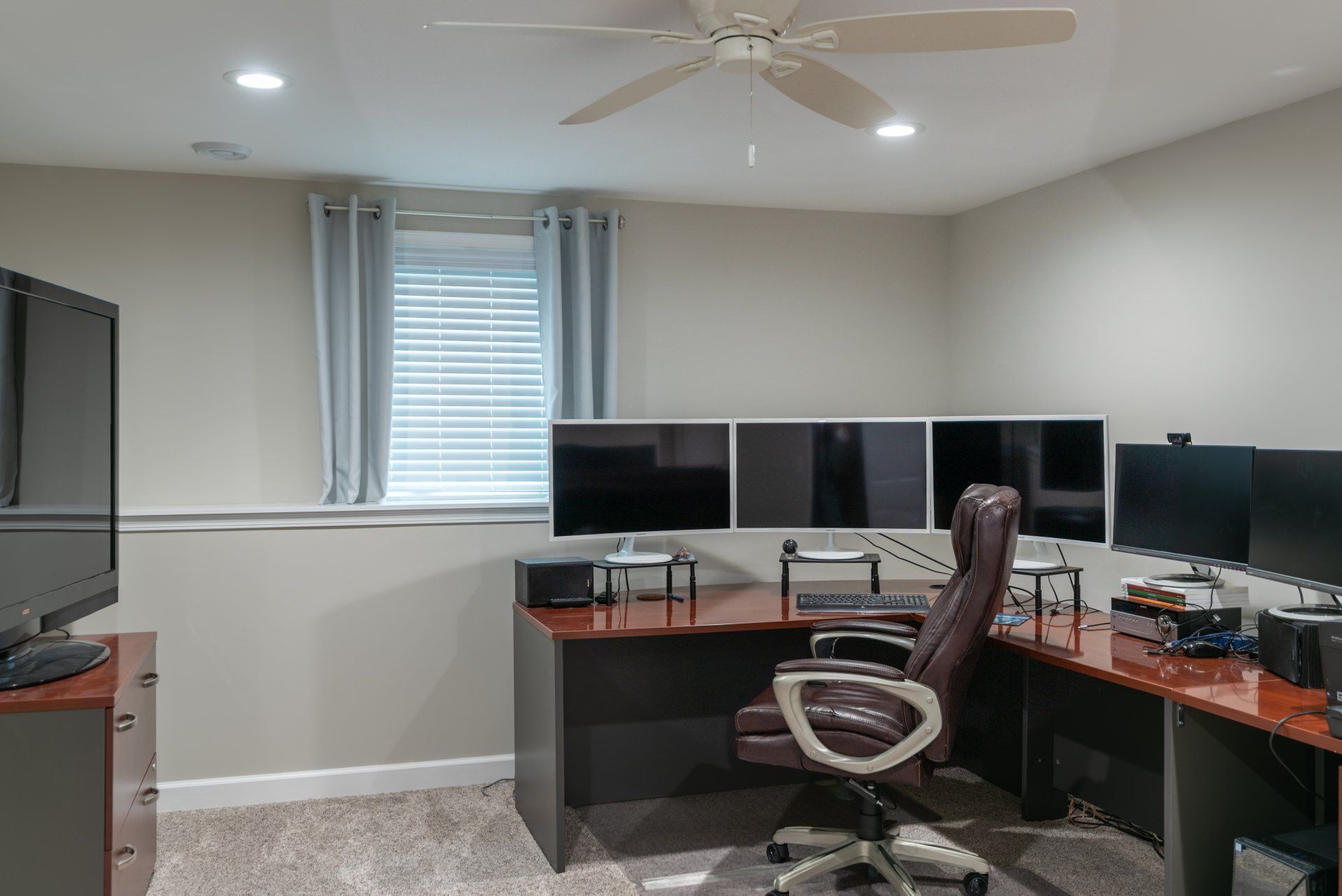A room with a desk , chair , computer monitors and a ceiling fan.