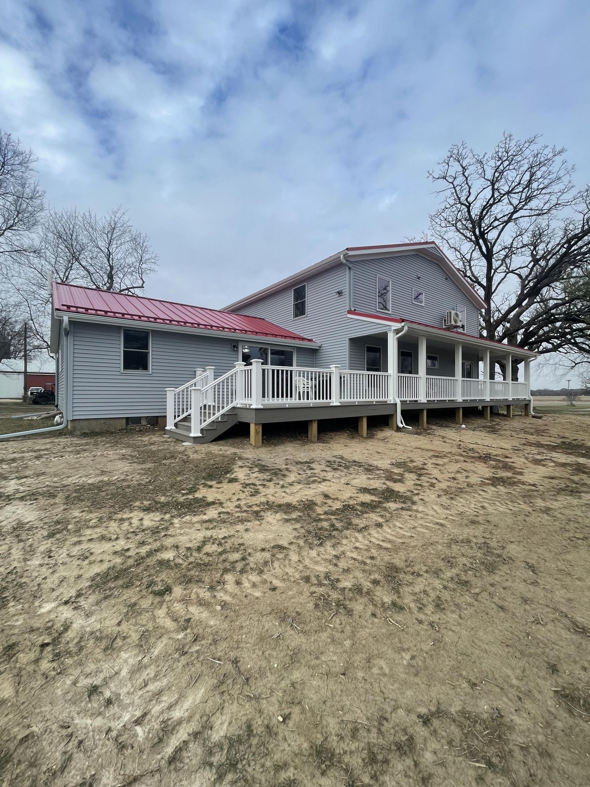 A white house with a red roof and a white deck is sitting on top of a dirt field.