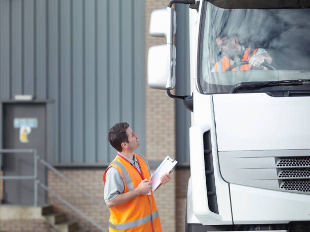 Man in orange vest inspecting a white truck with the driver visible in the cab. Gray building in the background.