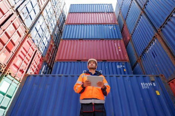 Worker in orange vest and hard hat with tablet, inspecting shipping containers at a port.