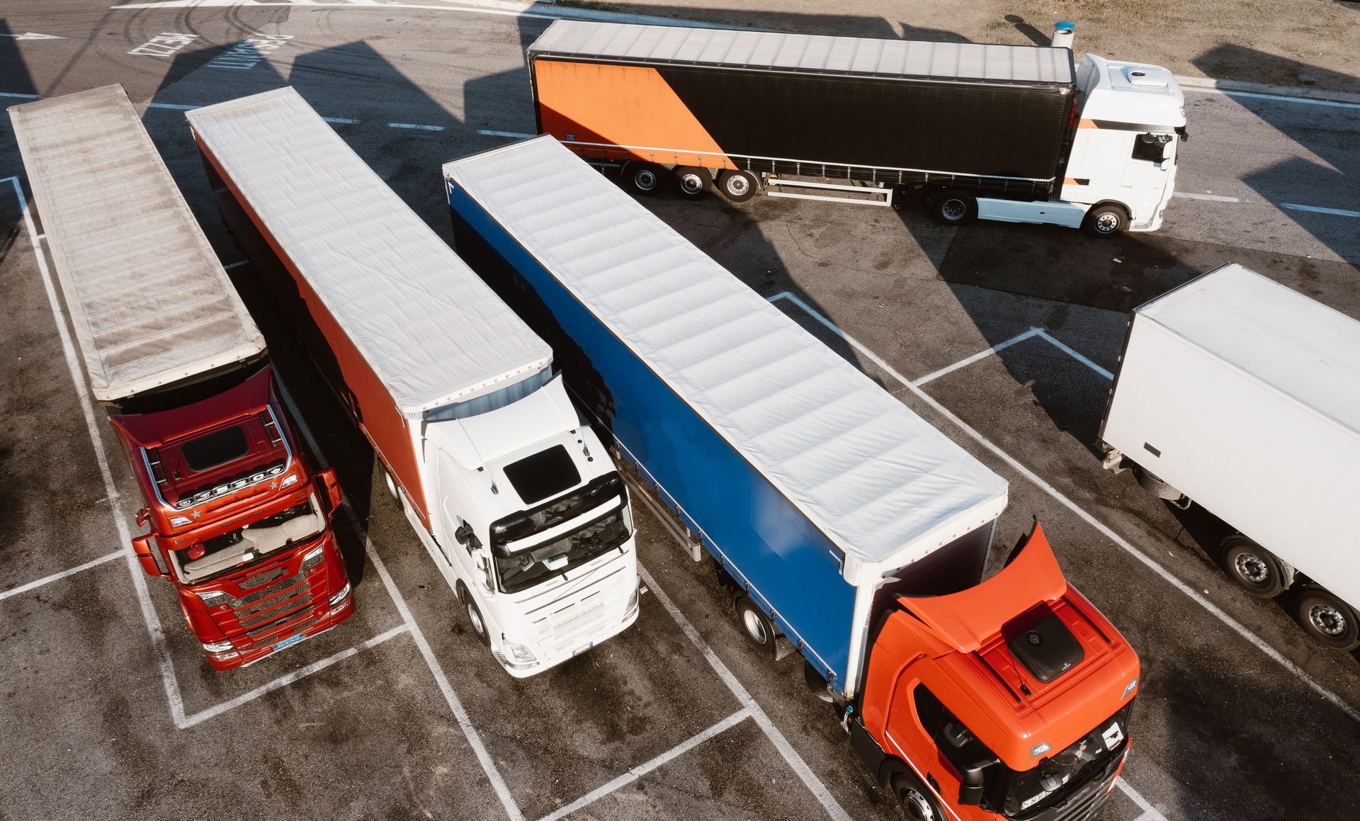 Overhead view of several semi-trucks parked in a lot; red, white, orange, and blue trailers.