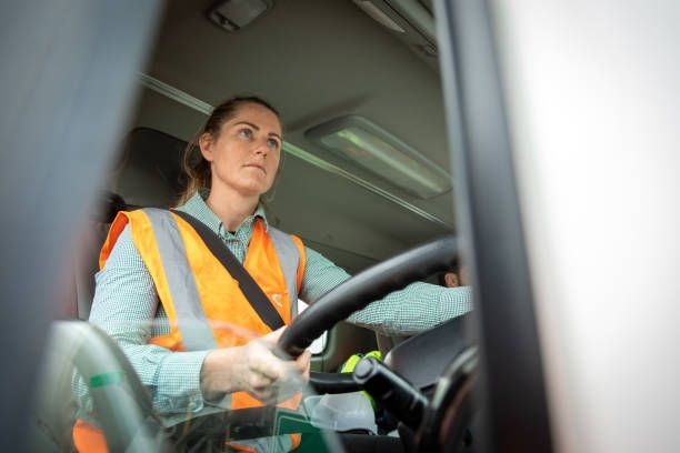 Woman in safety vest driving a vehicle, focused expression.