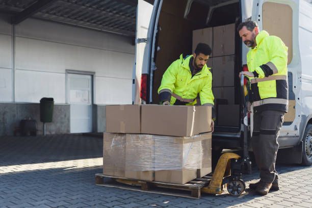 Two workers in yellow safety vests load boxes onto a van from a warehouse dock.