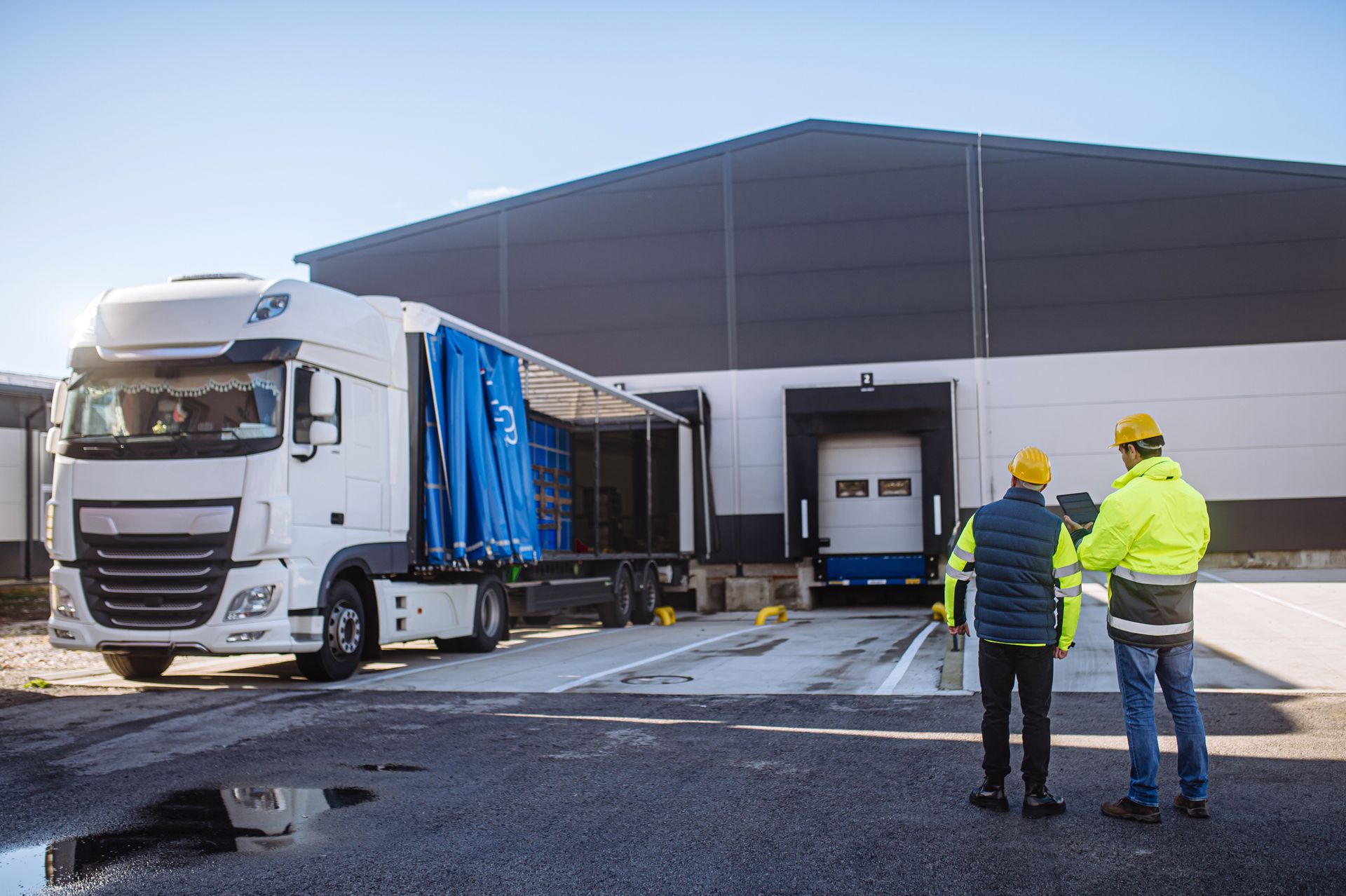 Truck unloading at a warehouse dock, two workers in safety gear observing.