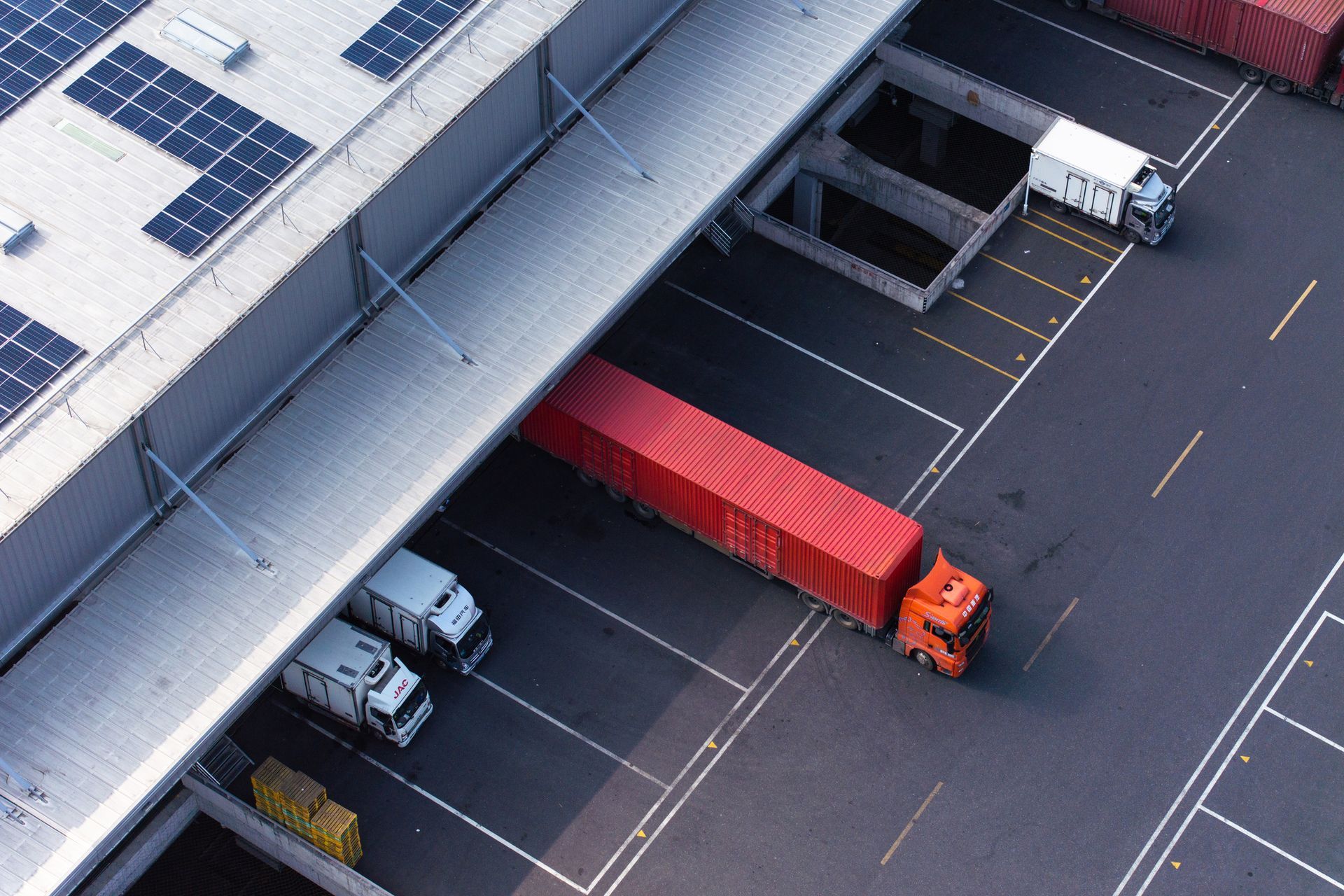 Truck loading at a warehouse dock, red container, white trucks, solar panels on roof.