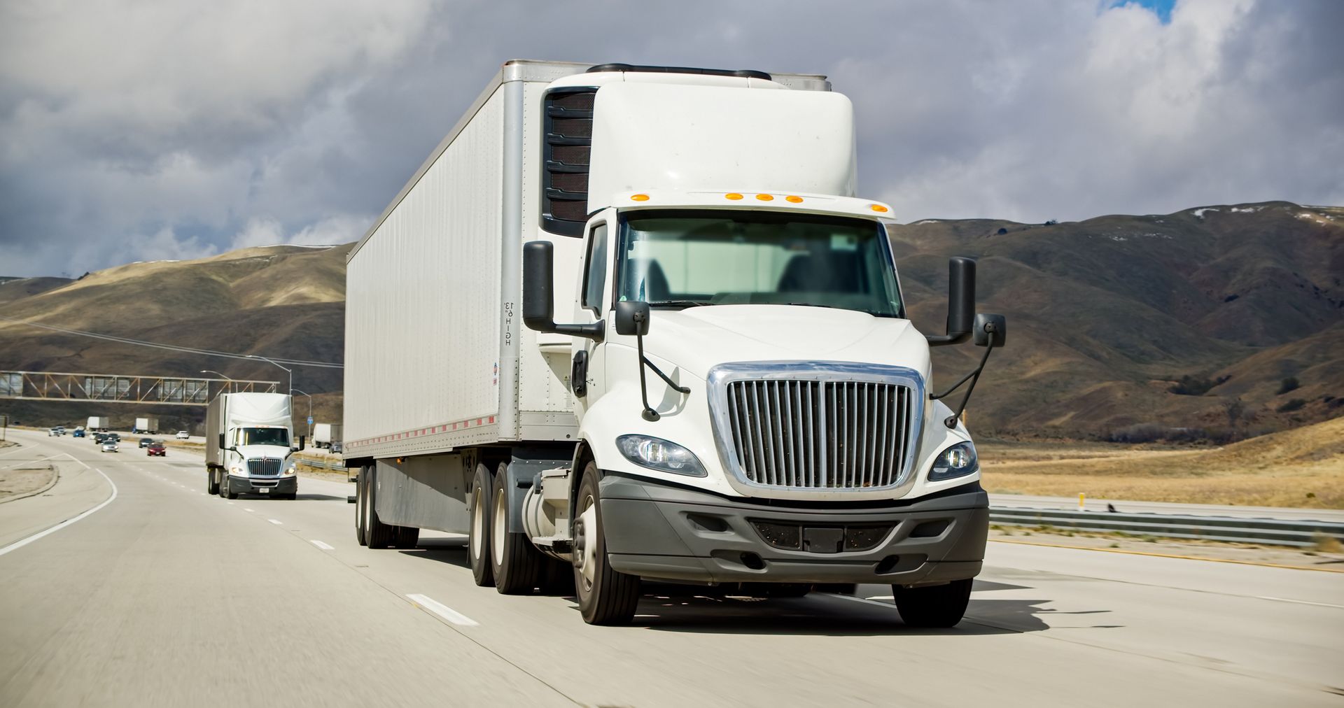 White semi-truck driving on a highway under a cloudy sky, with another truck in the distance and hills in the background. White semi-truck driving on a highway under a cloudy sky, with another truck in the distance and hills in the background.