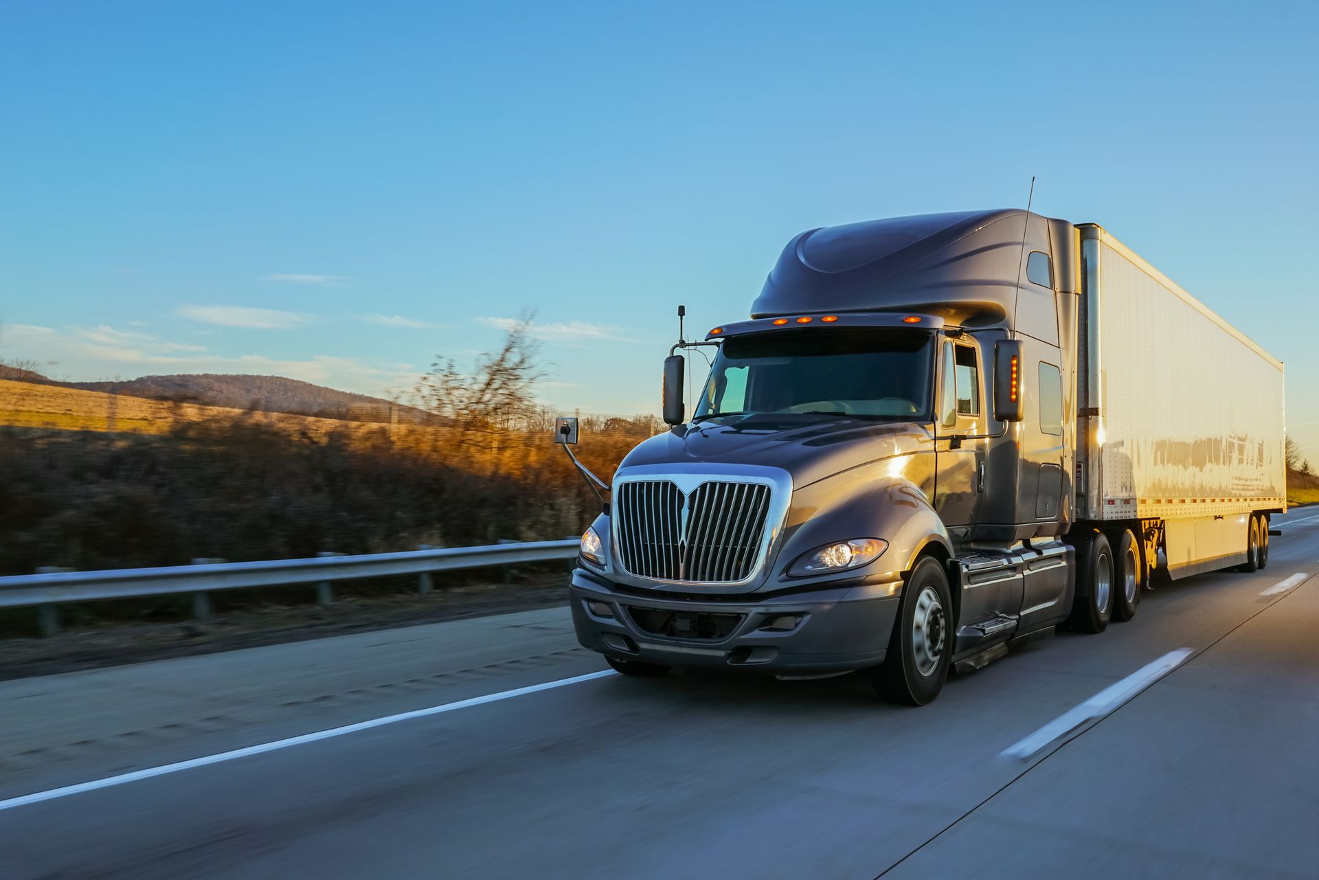 Semi-truck driving on a highway at sunset; gray truck, white trailer, blue sky.
