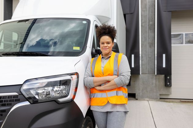 Woman in safety vest stands by a white delivery van at a loading dock, arms crossed, smiling.