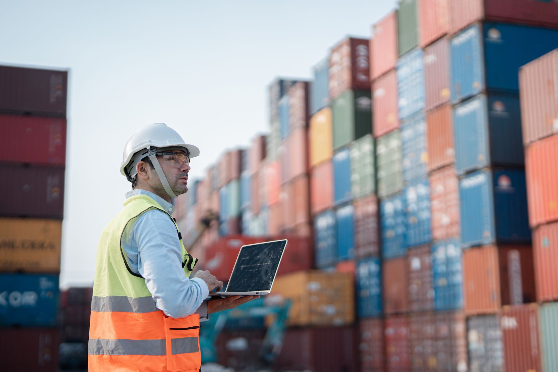 Man in safety gear with laptop, inspecting shipping containers at a port.