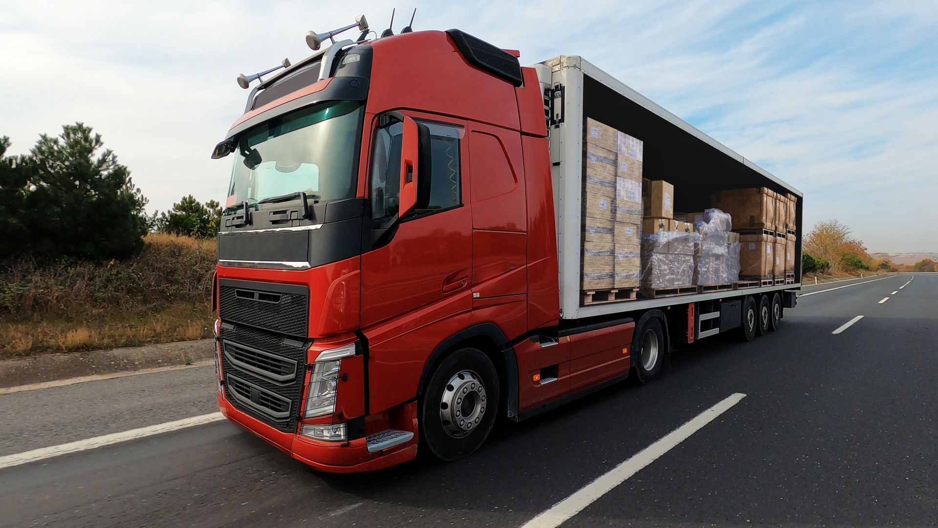 Red semi-truck on a highway, carrying cargo in an open trailer.