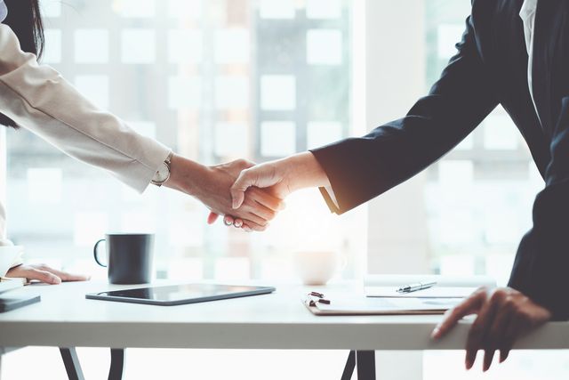Two people in business attire shaking hands across a bright office desk