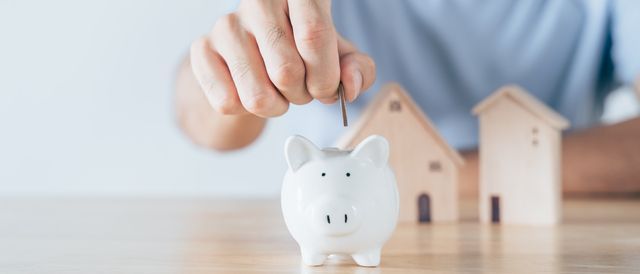 Hand placing a coin into a white piggy bank beside two small wooden houses