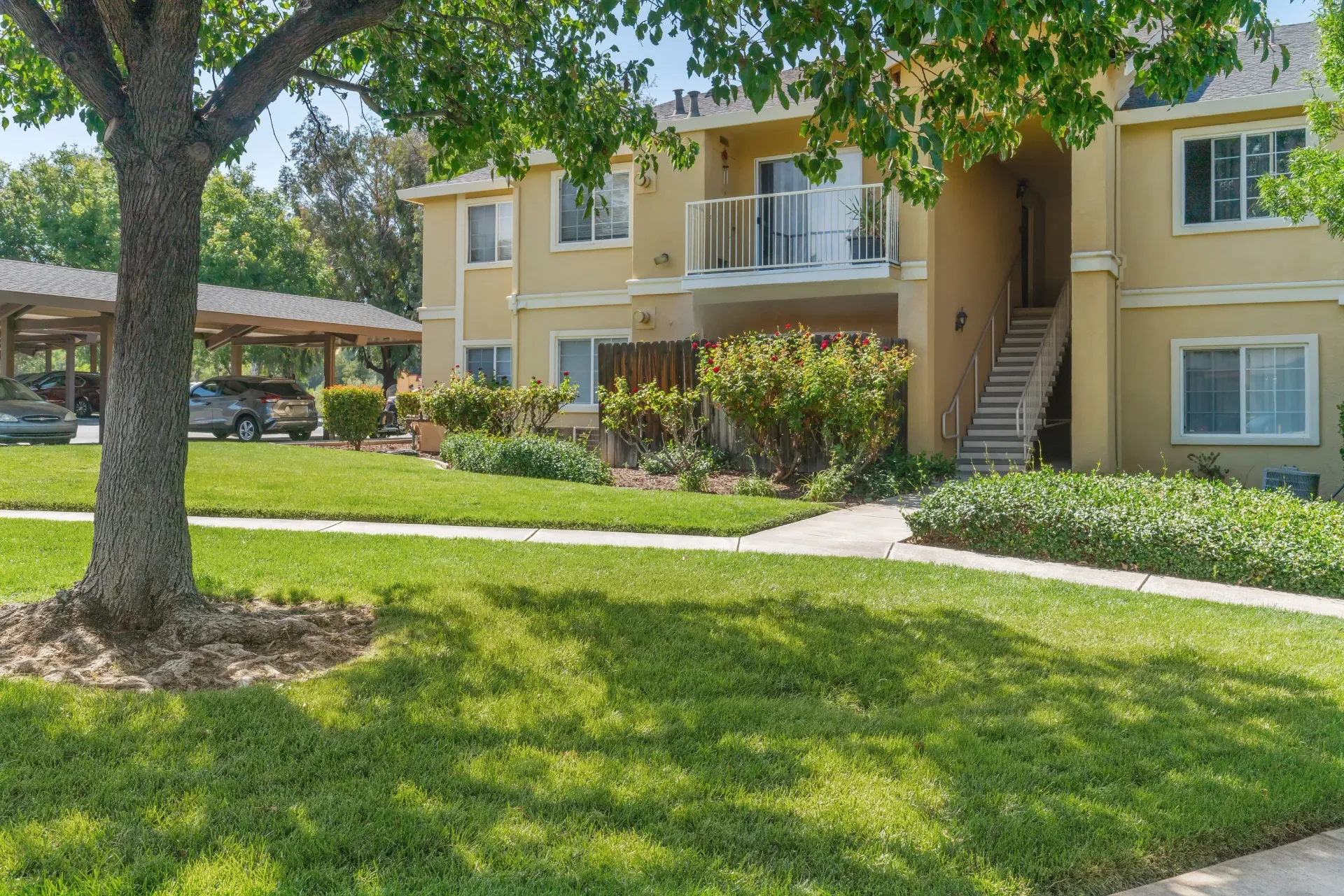 Beige two-story apartment building with stairs to upper units, surrounded by a green lawn and trees.