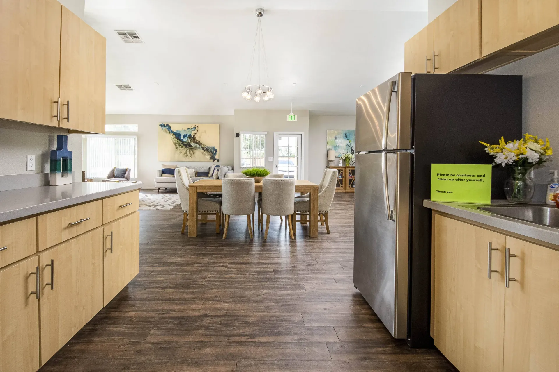 Open-concept apartment kitchen with light wood cabinets, gray countertops, stainless steel fridge, and a dining table nearby.