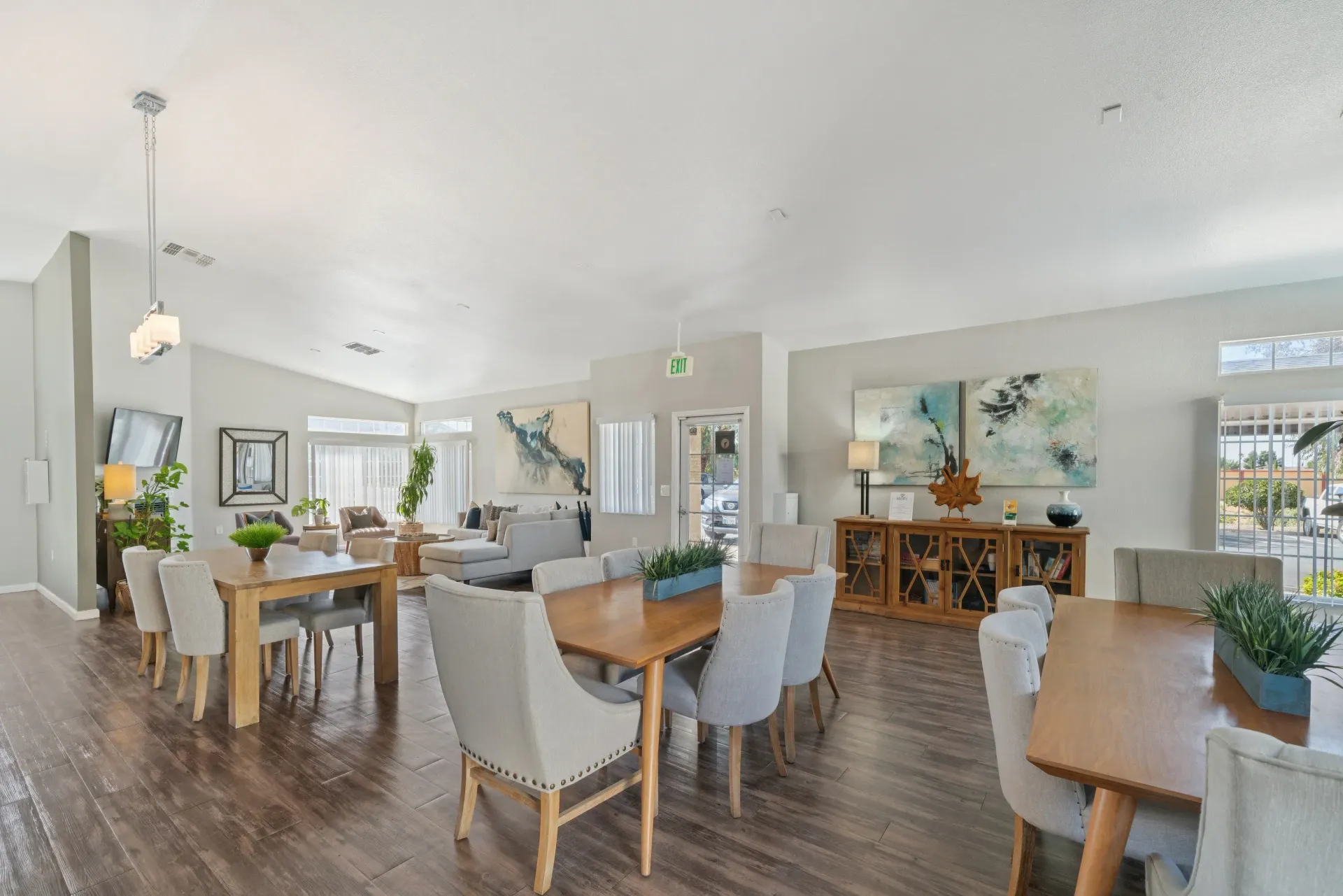 Bright communal lounge with multiple dining tables, gray upholstered chairs, plants, and artwork.