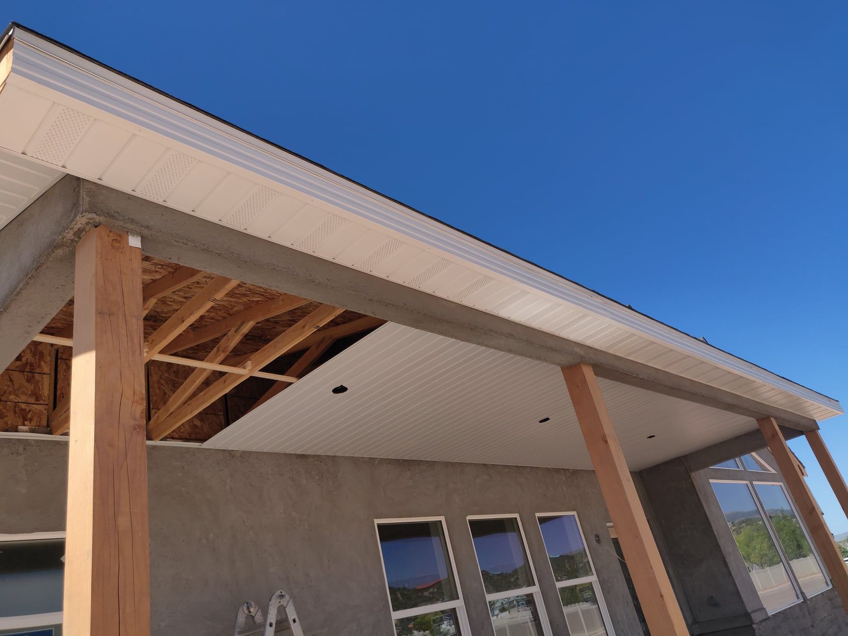 Construction of a porch with wooden posts, stucco wall, and white ceiling panels under a blue sky.