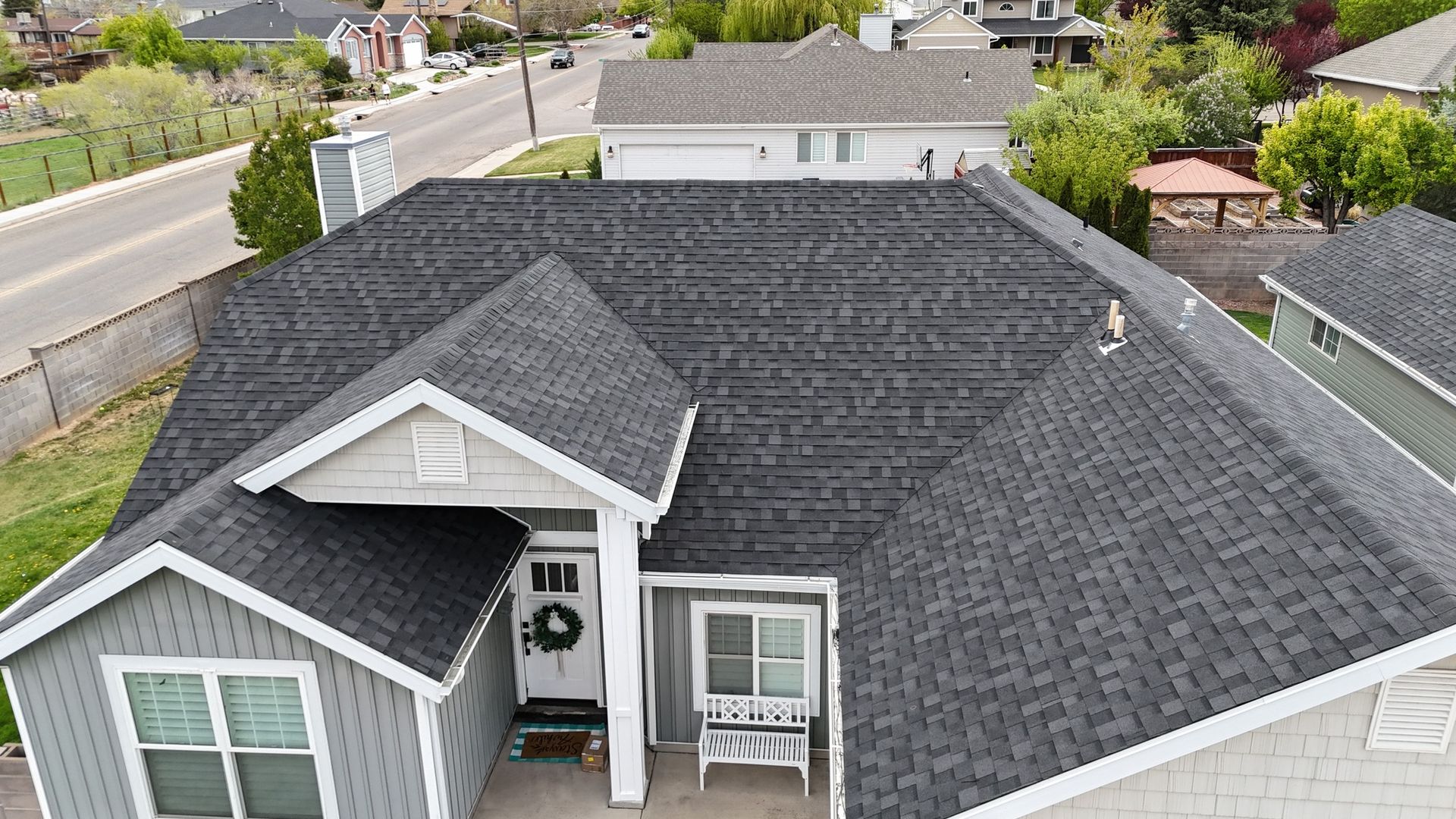 House with dark gray asphalt shingle roof. Gray siding, white trim, front door, and surrounding greenery.