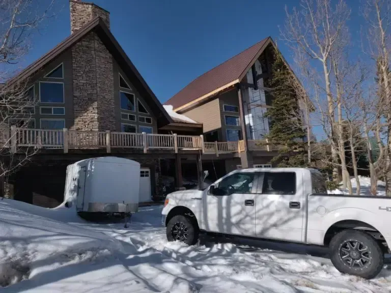 White truck and trailer parked in snow in front of a large, multi-gabled house with brown stone and siding.