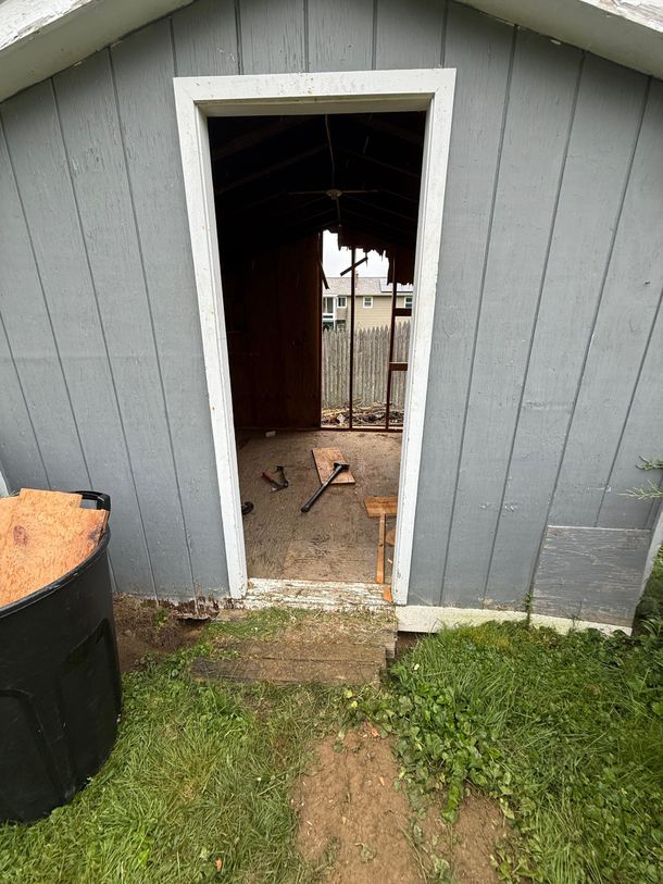 Gray shed with open doorway, showing a dark interior and a dirt path leading to it from the grass.