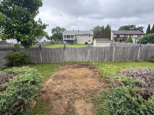 Backyard garden with a dirt path, shrubs, and a wooden fence, with houses in the background