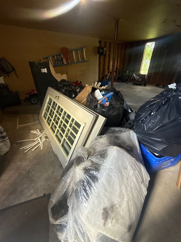 Cluttered room with a broken window frame, covered furniture, and trash bags on the floor