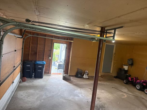 Empty garage interior with concrete floor, exposed ceiling beams, and bins near an open door.