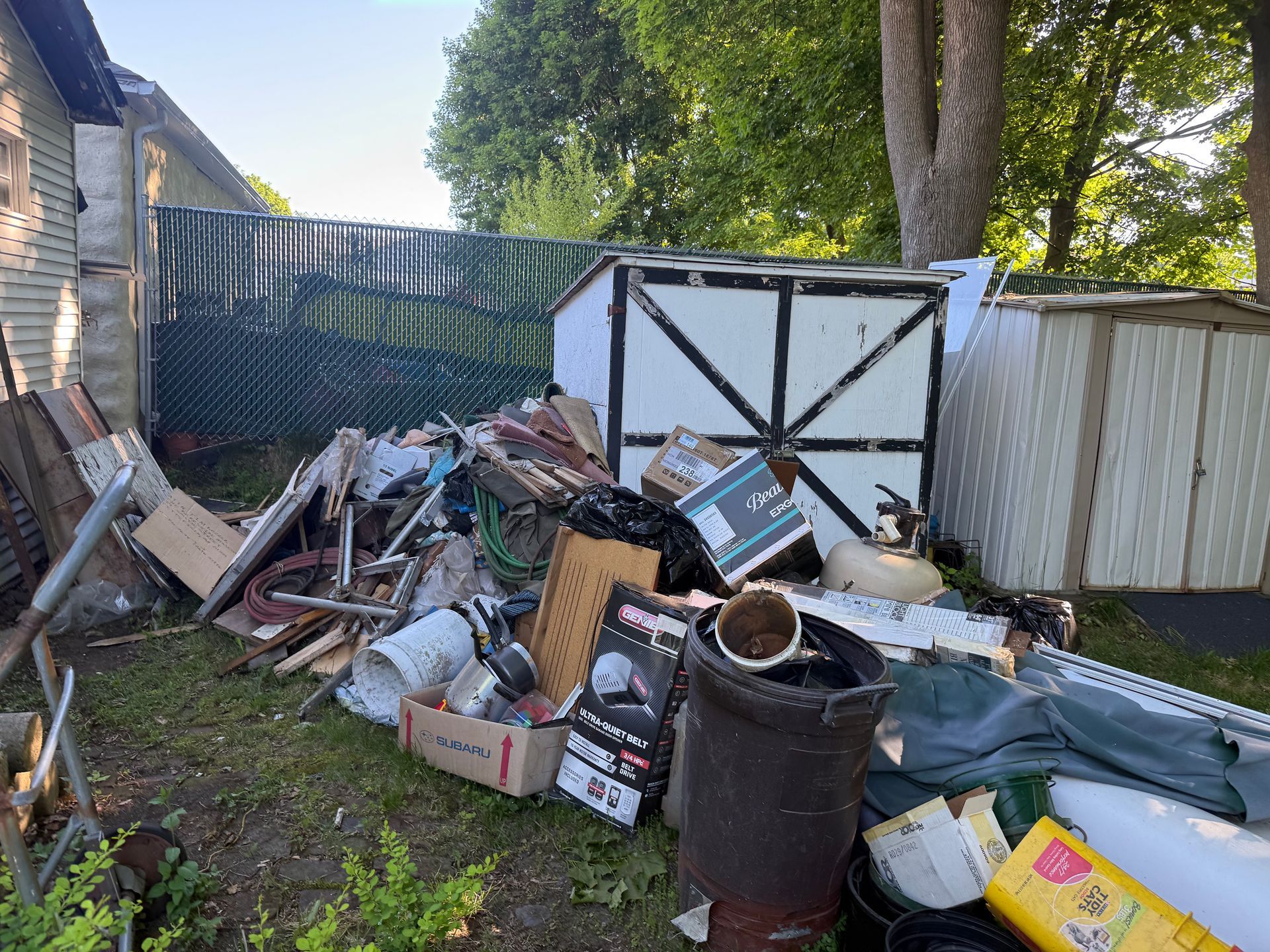 Piled junk and debris in a fenced yard beside a shed and trees