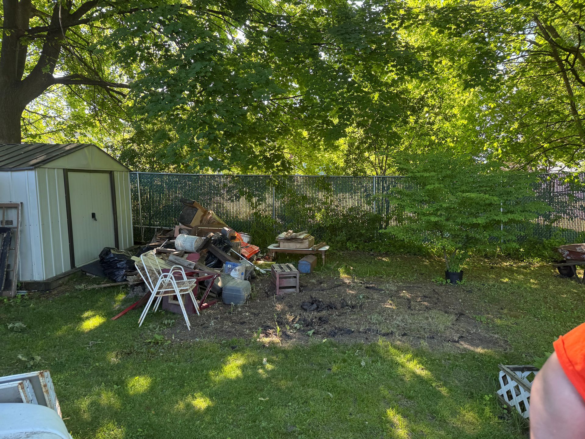 Backyard with shed, scattered clutter, and a wooden fence under leafy trees.