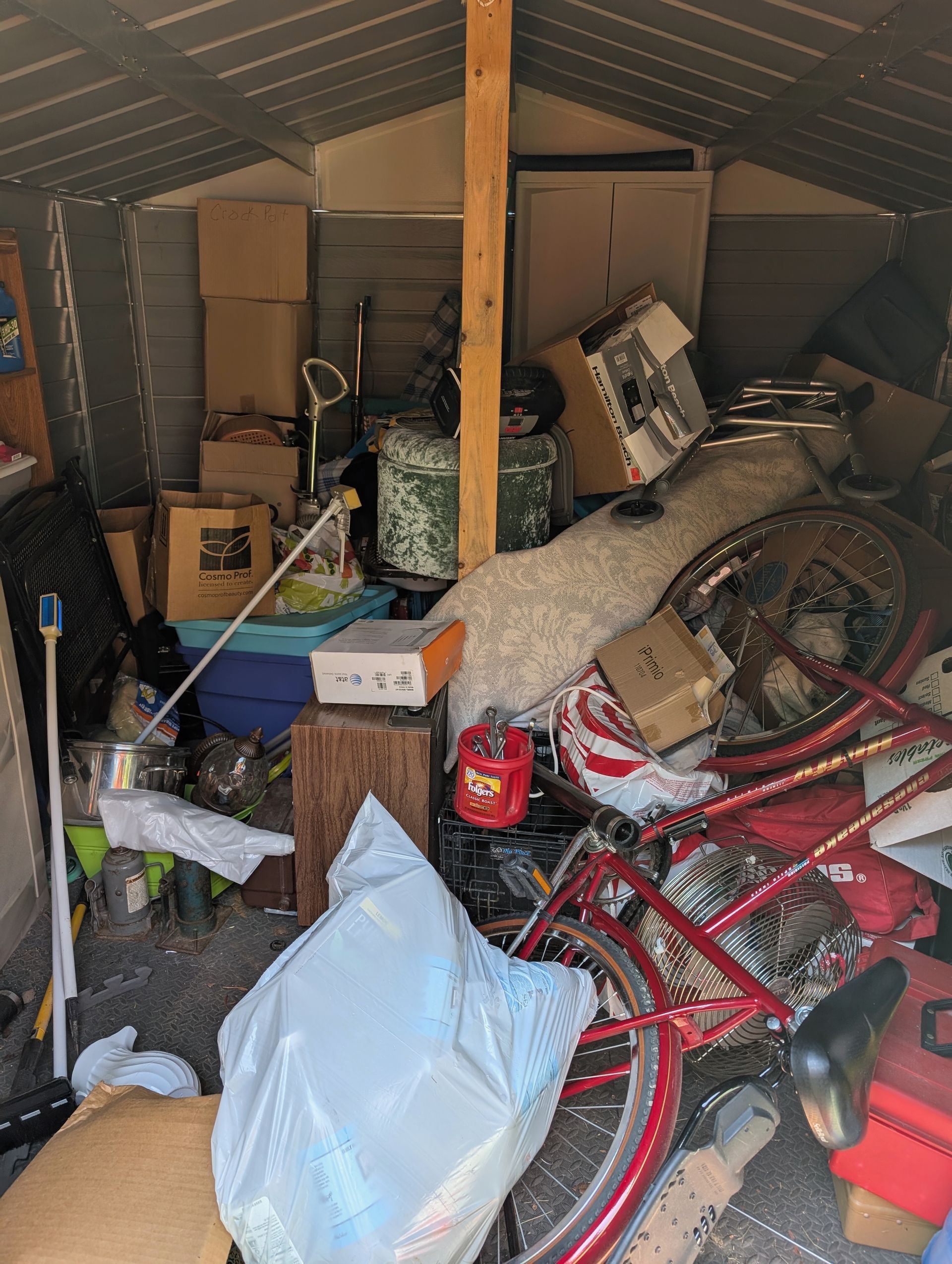 Cluttered storage shed packed with boxes, bags, tools, and bicycles under a slanted roof.