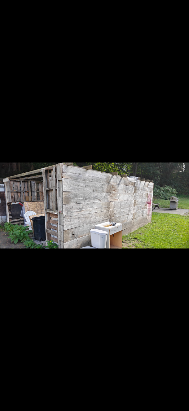 Small stone shed with a wooden door in a grassy yard, with buckets and a barrel beside it.