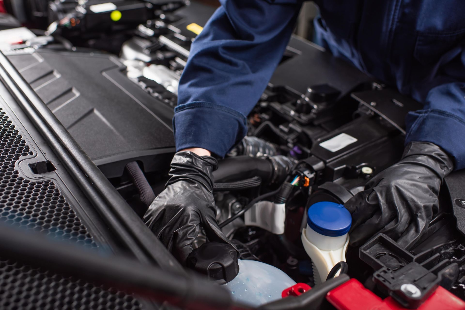 Mechanic in blue uniform and black gloves working on a car engine.