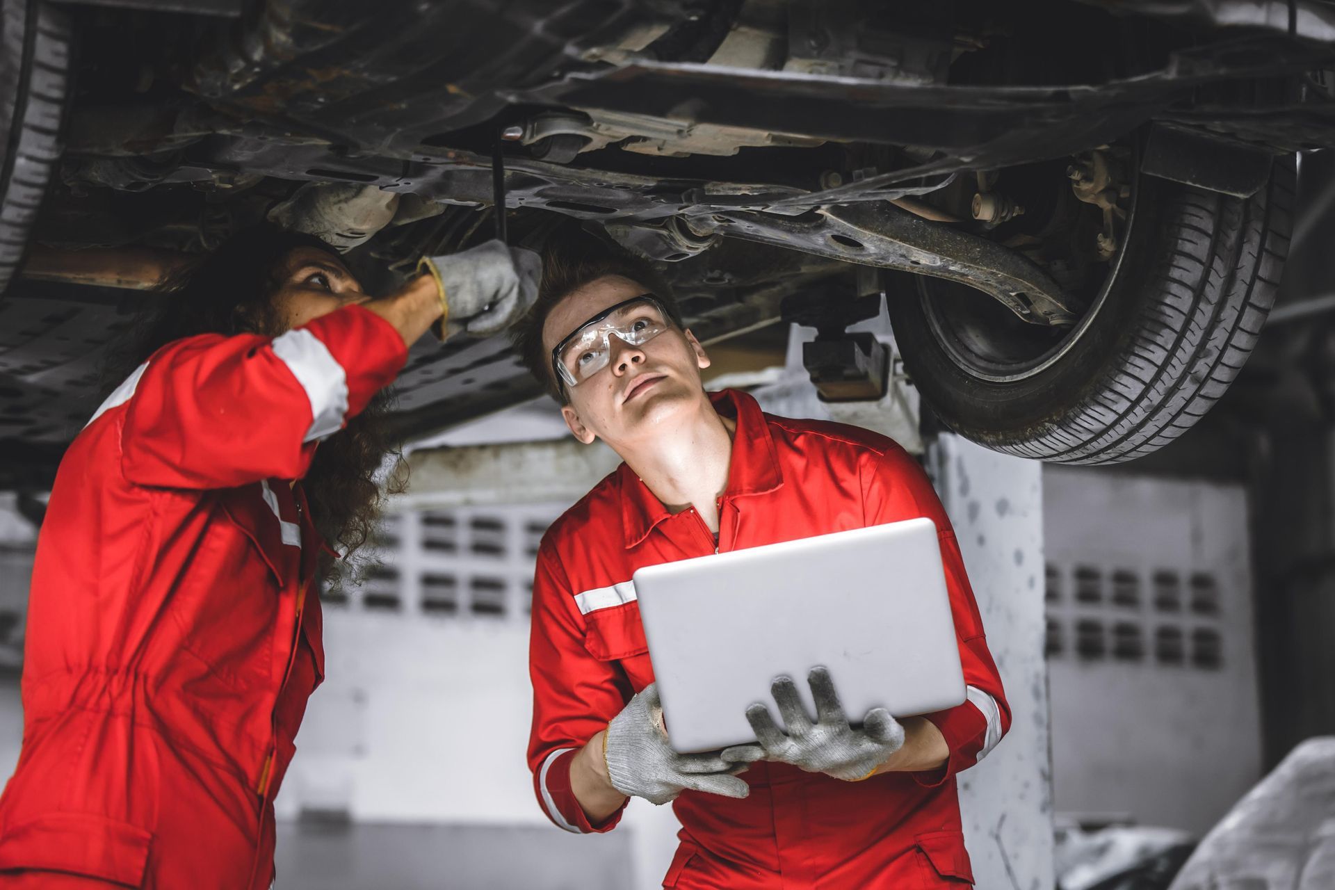 Two mechanics in red jumpsuits examine a car from below, one using a laptop.