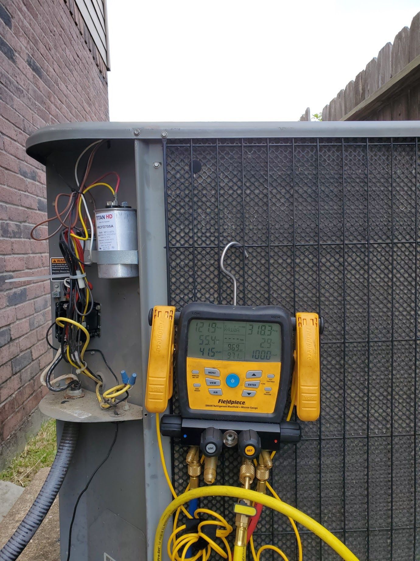 HVAC unit with gauges attached, measuring pressure. Yellow and black tool is center of focus. Wires and capacitor visible.
